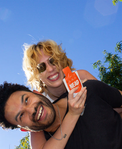 Two people outdoors with a clear blue sky, dynamically holding holding a Peach NEAP protein gel pouch.